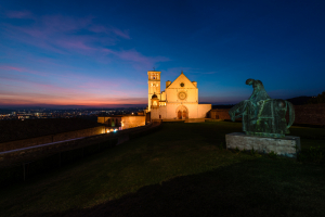 Die beleuchtete Basilika des Hl. Franziskus von Assisi und die bronzene Reiterstatue im Zwielicht der Abenddämmerung, Umbrien, Italien